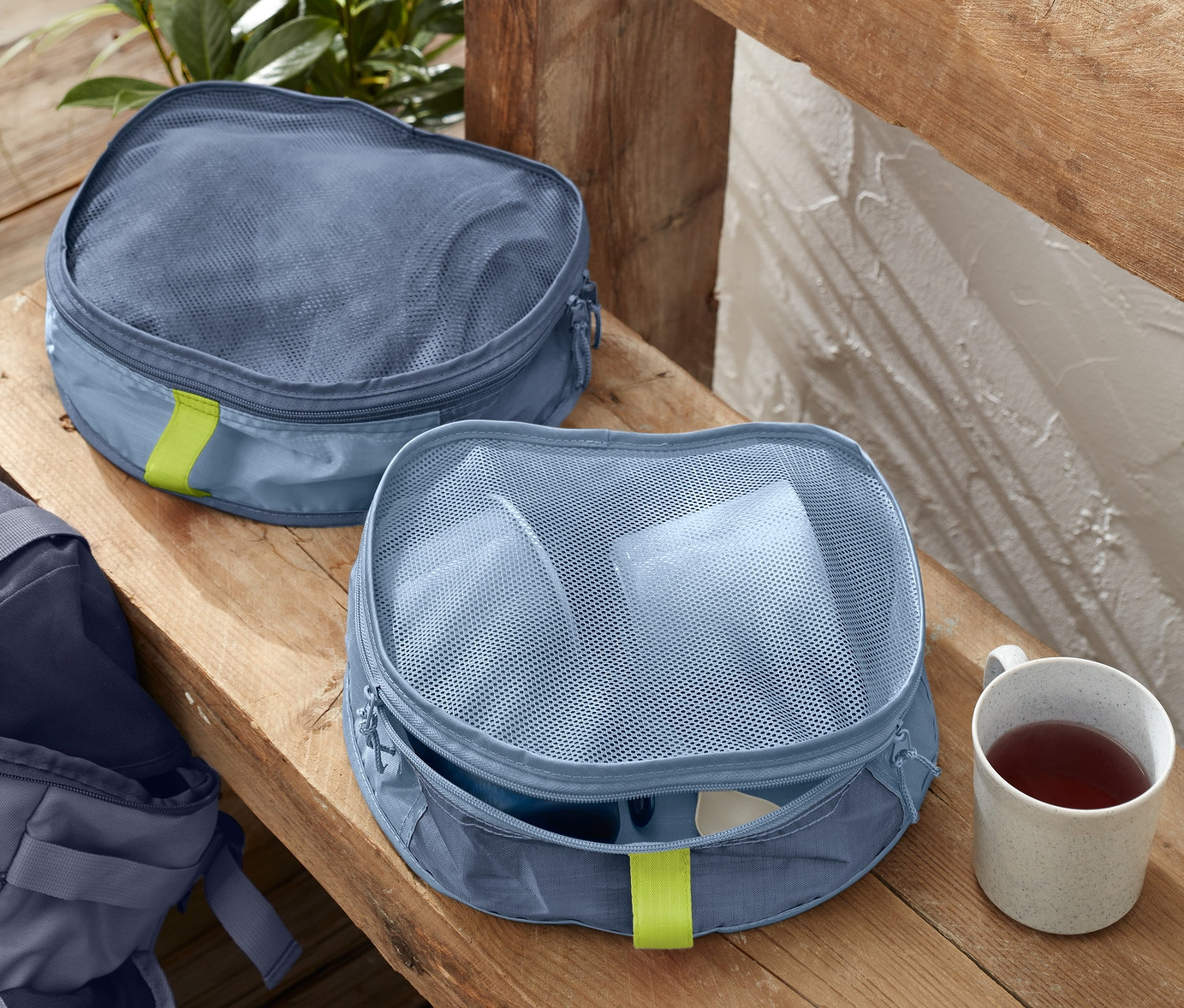 Dwa organizery do plecaka leżą na drewnianej ławce obok filiżanki. W tle widać plecak.