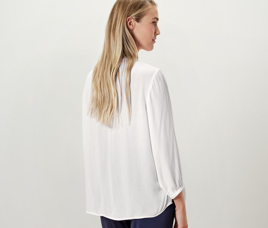 Blondynka pozuje tyłem do kamery, ubrana w białą bluzkę-tunikę.