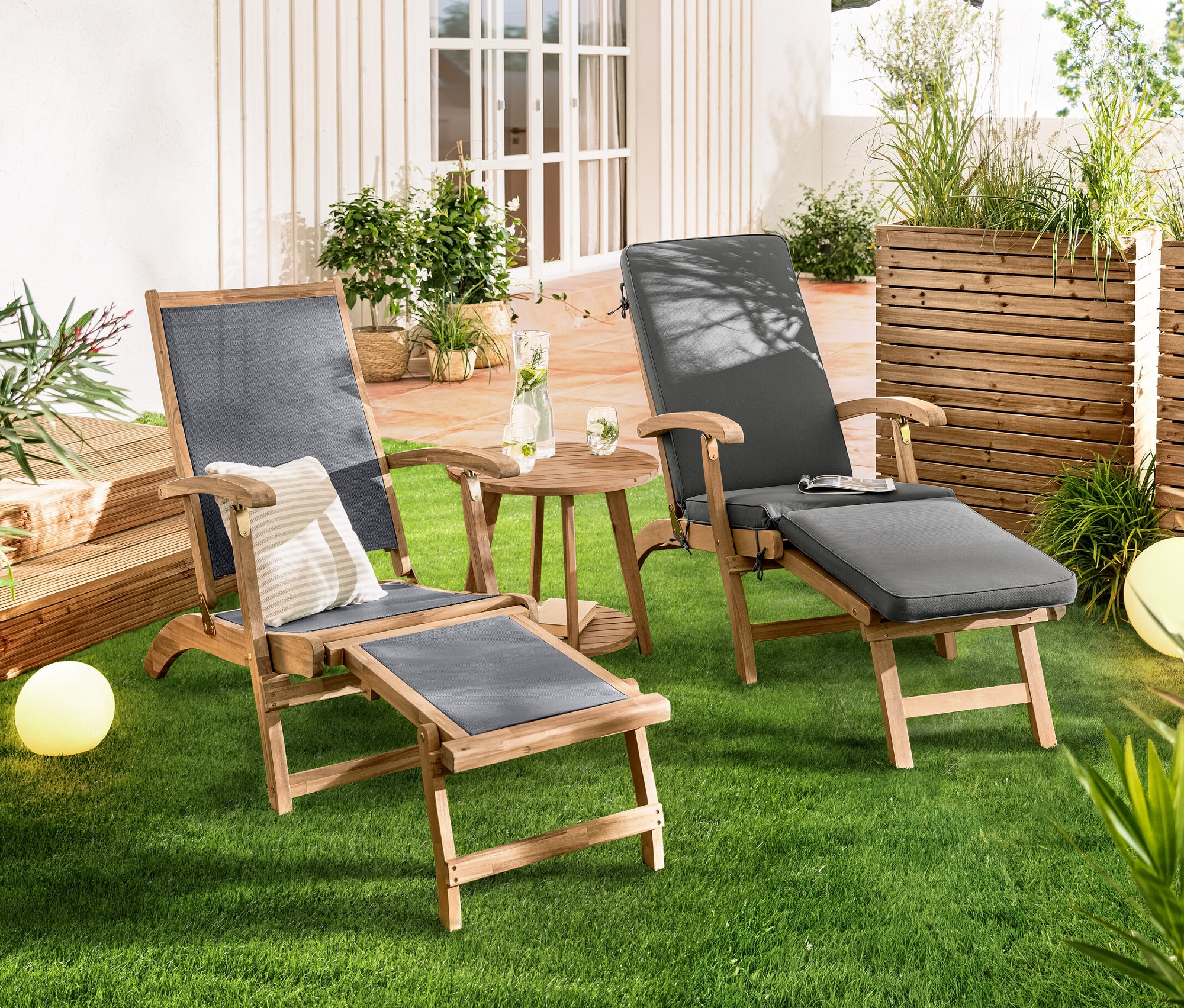 Dwa leżaki „Lenja” z wysuwanym podnóżkiem stoją obok siebie na trawniku. Pomiędzy nimi znajduje się stolik dwupoziomowy „Lenja”. W tle widoczne są lampy solarne w kształcie kuli, w kolorze piasku, ok. 20 cm i lampy solarne w kształcie kuli, w kolorze piasku, ok. 30 cm.