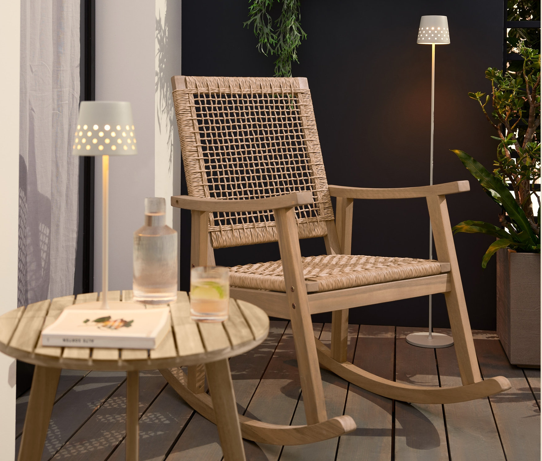 Na tarasie stoi fotel bujany z plecionką polirattanową, dwupoziomowy stolik „Lenja” z imitacją drewna tekowego, metalowa lampa stołowa LED z możliwością ładowania oraz metalowa lampa podłogowa LED z możliwością ładowania.