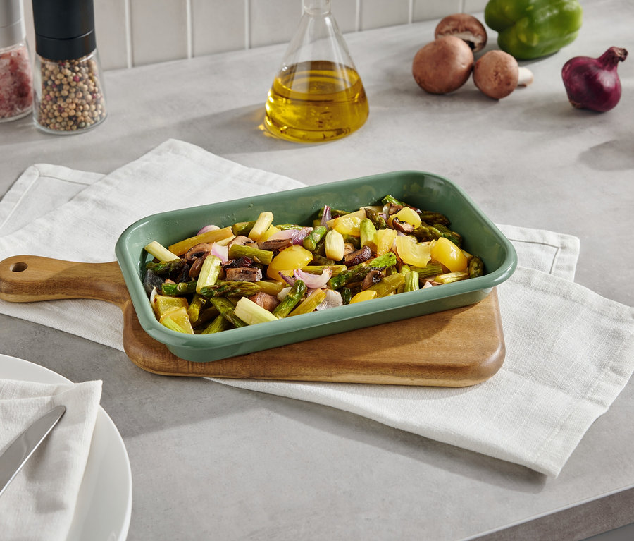 Zielone naczynie do zapiekania z pieczonymi warzywami na desce z serwetką. W tle grzyby, papryka, cebula i przyprawy.