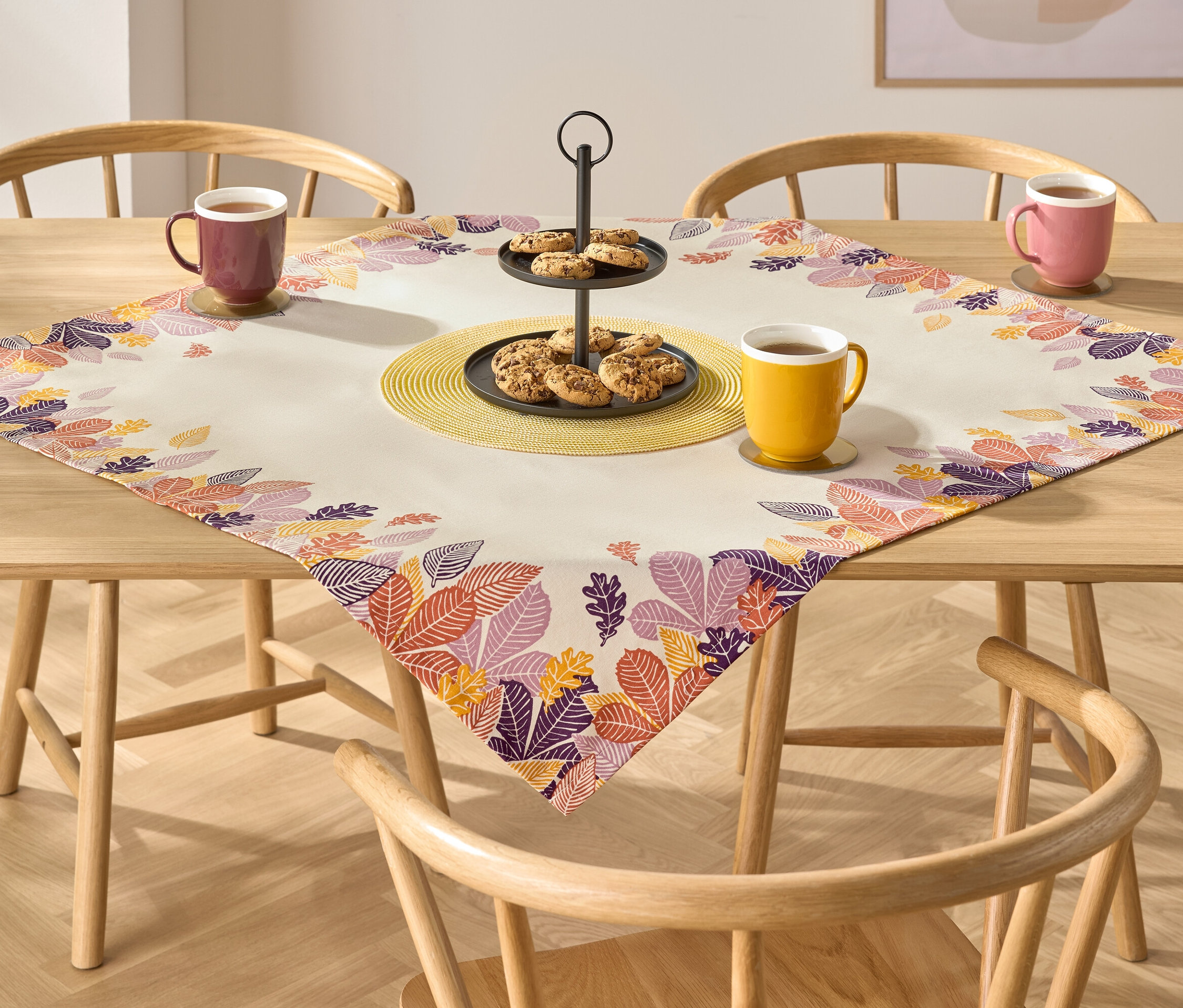 Na drewnianym stole leży obrus z jesiennymi motywami liści. Na obrusie stoi etażerka z ciasteczkami i trzy kubki z napojami.