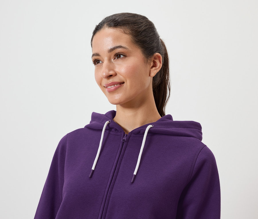 Kobieta z brązowymi włosami związanymi w kucyk ma na sobie fioletową bluzę z kapturem zapinaną na zamek.