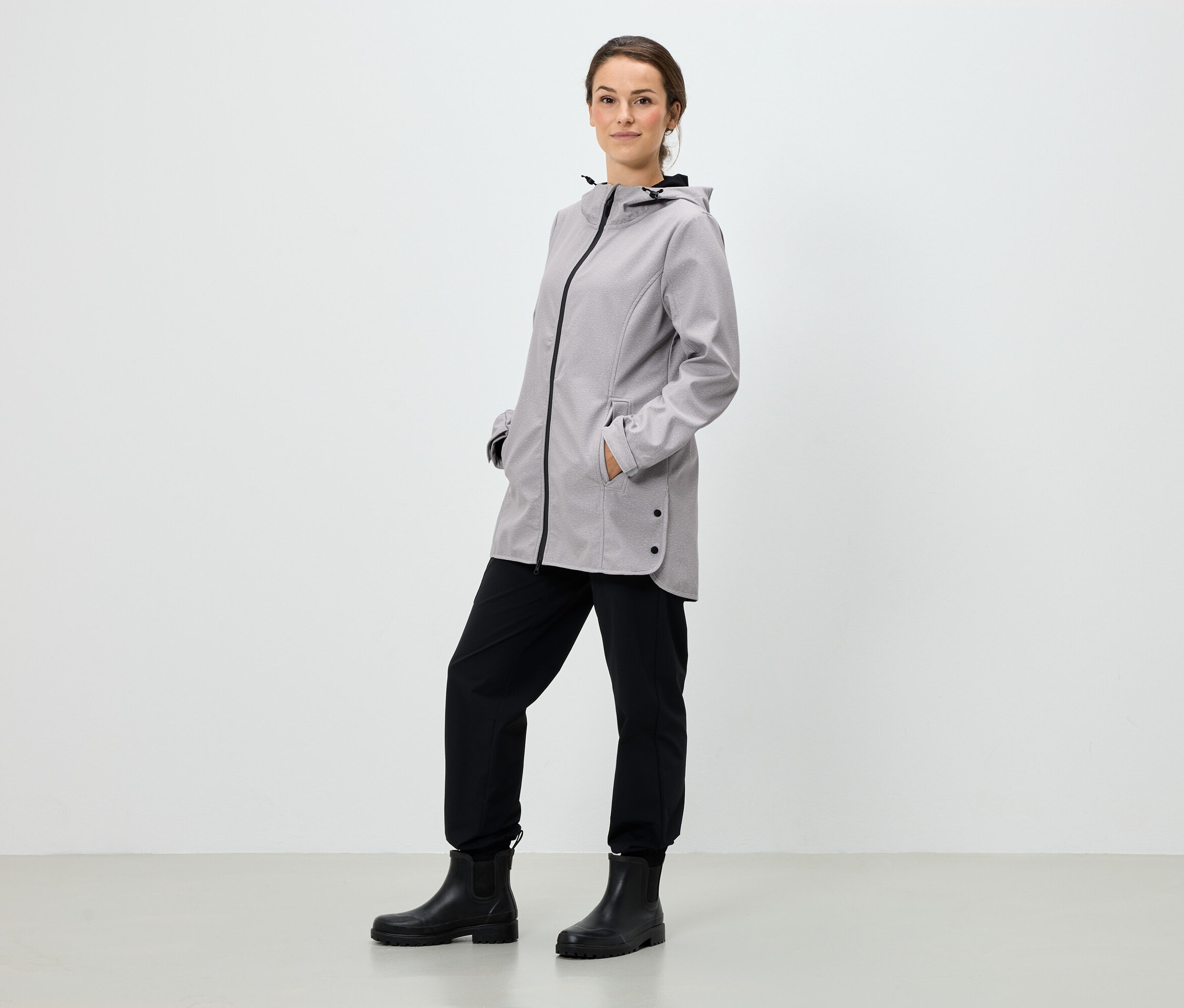 Kobieta stoi z rękami w kieszeniach szarej kurtki z kapturem, czarnych spodniach i czarnych butach.