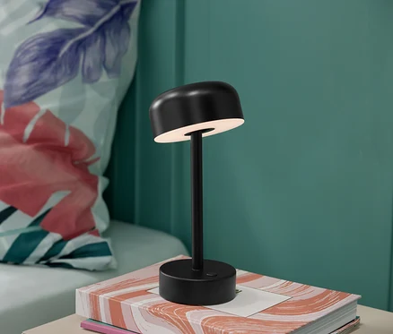 Czarna akumulatorowa lampa stołowa LED stoi na książce, w tle poduszka w kwiaty.