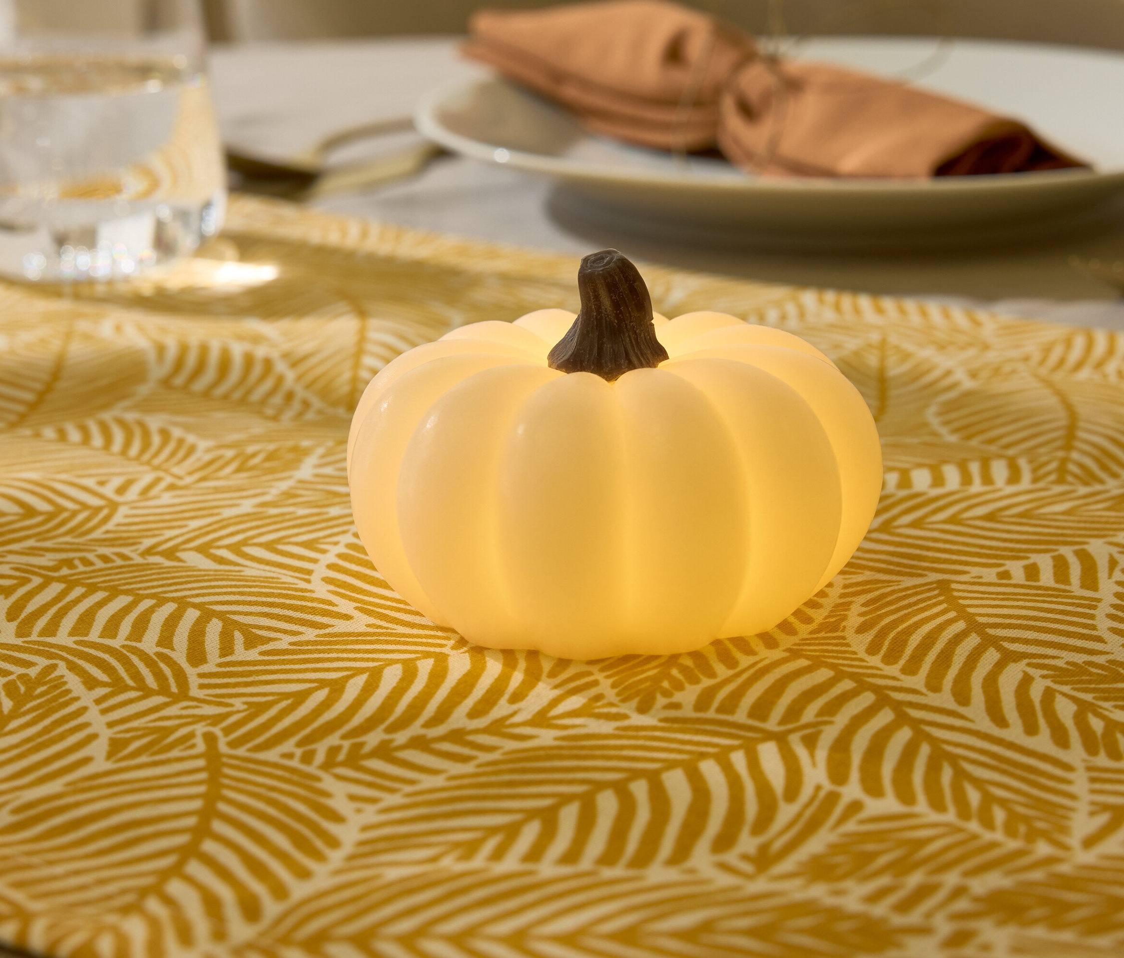 Dekoracyjna lampka w kształcie dyni na bieżniku ze wzorem liści, w tle szklanka wody i talerz.