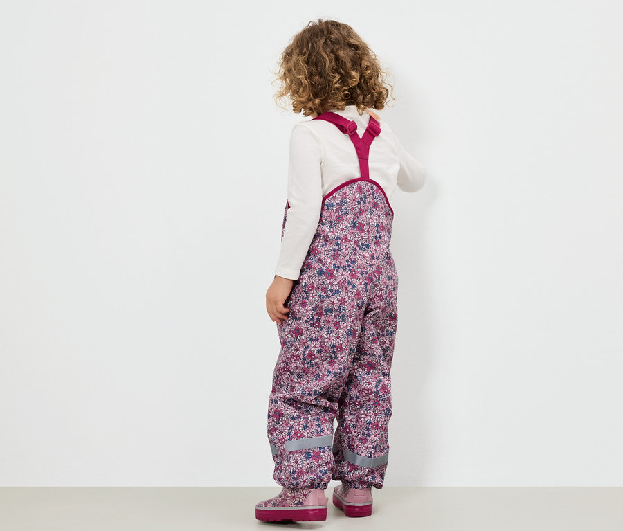 Dziewczynka w kwiecistych spodniach na szelkach i różowych butach stoi tyłem do widza.