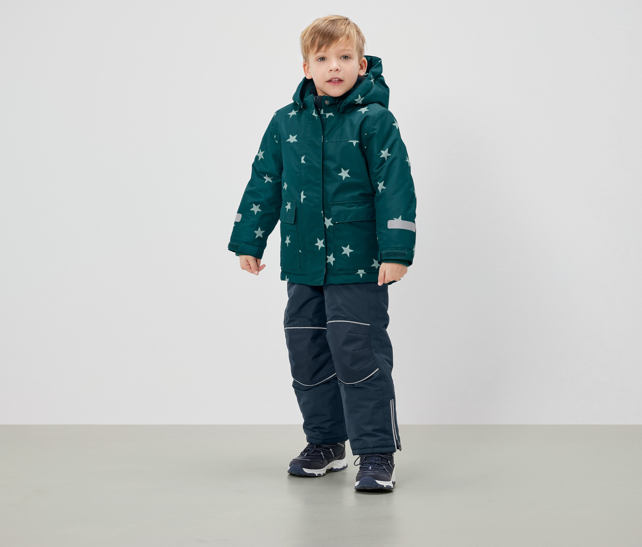 Chłopiec stoi w zielonej kurtce zimowej w gwiazdy i granatowych spodniach.