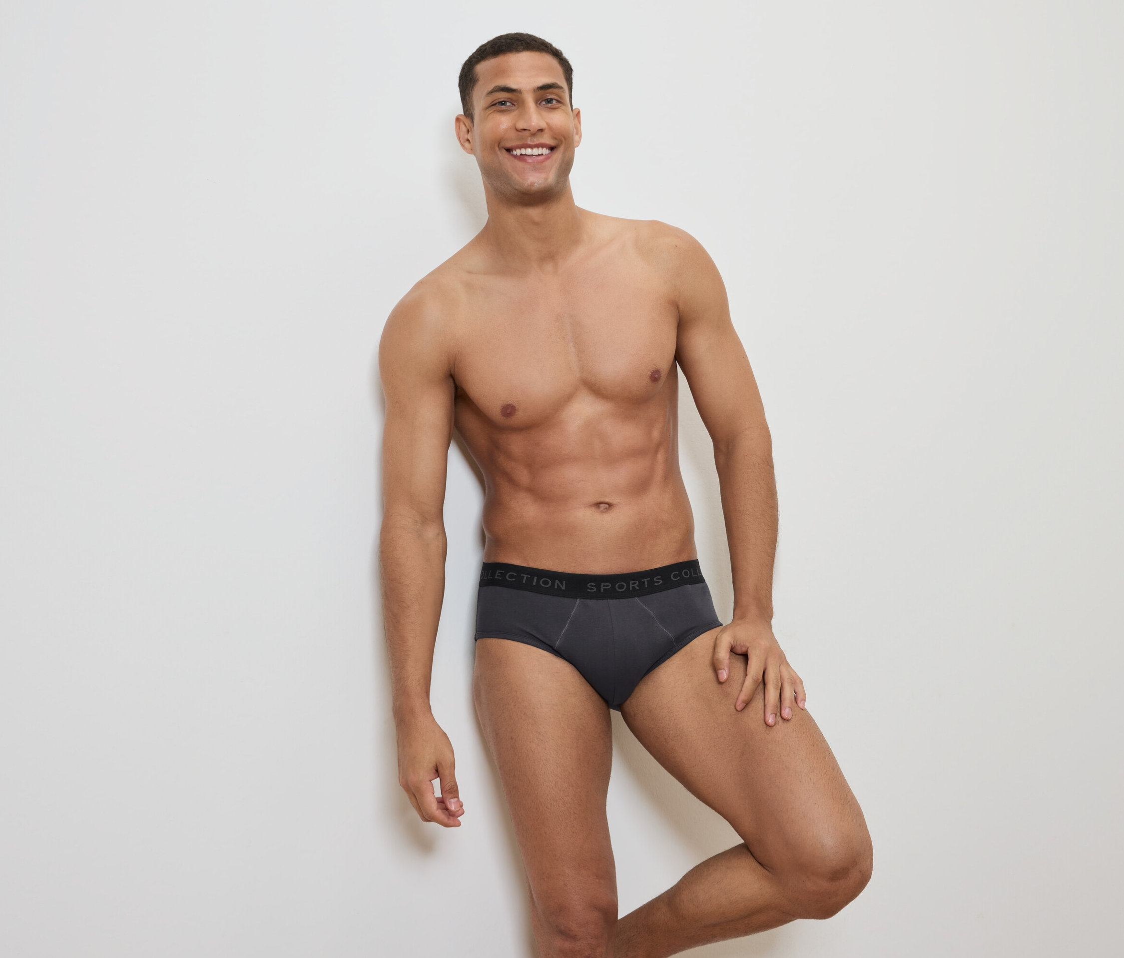 Uśmiechnięty mężczyzna z odsłoniętą górną częścią ciała pozuje w szarych slipach z czarnym pasem.