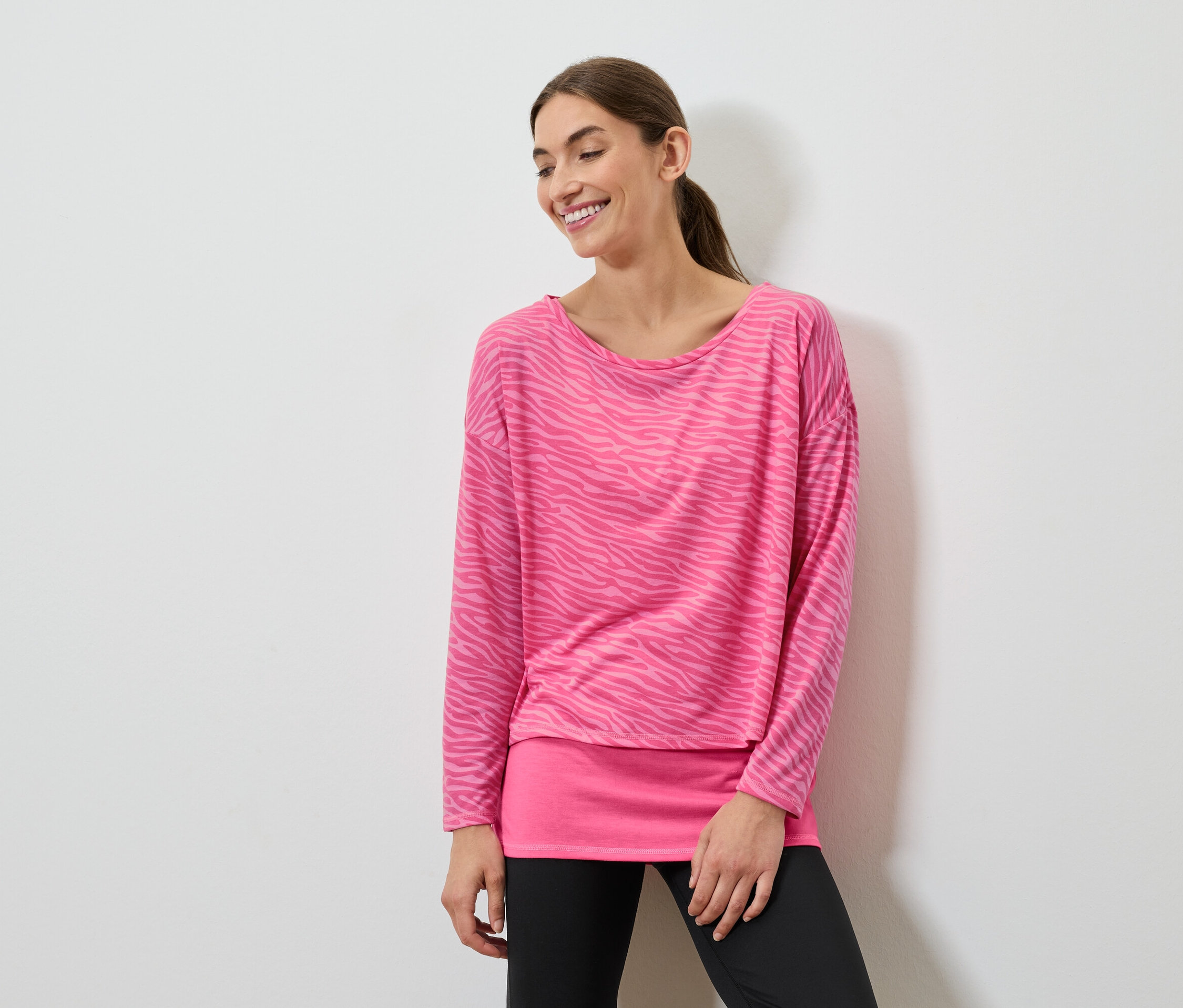 Uśmiechnięta kobieta pozuje w różowej bluzce z długim rękawem i czarnych legginsach.