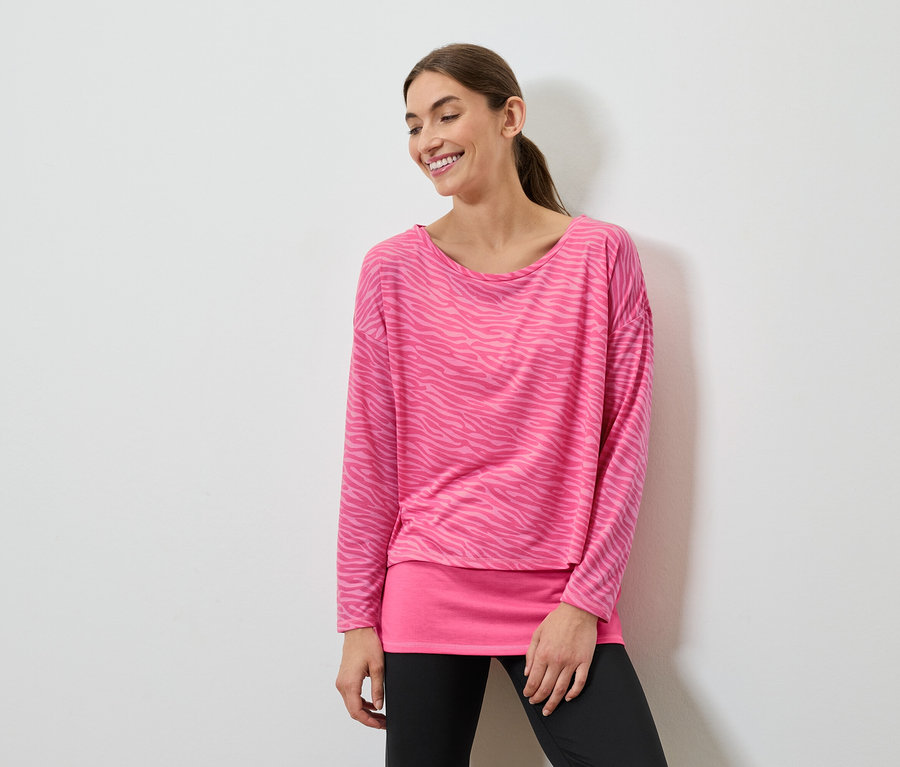 Uśmiechnięta kobieta pozuje w różowej bluzce z długim rękawem i czarnych legginsach.