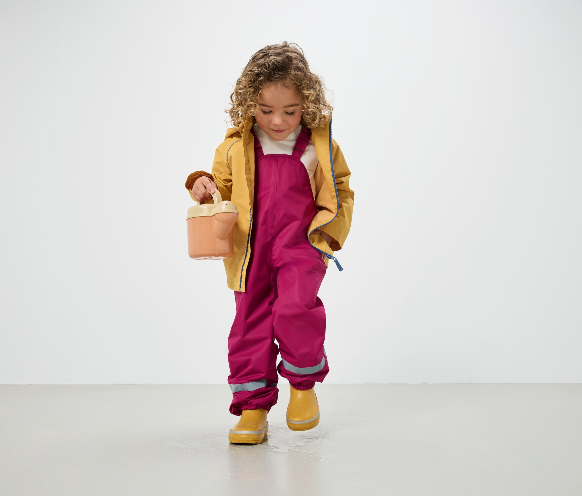 Dziecko z kręconymi włosami w żółtej kurtce, różowych spodniach i żółtych kaloszach trzyma konewkę i stoi na podłodze.