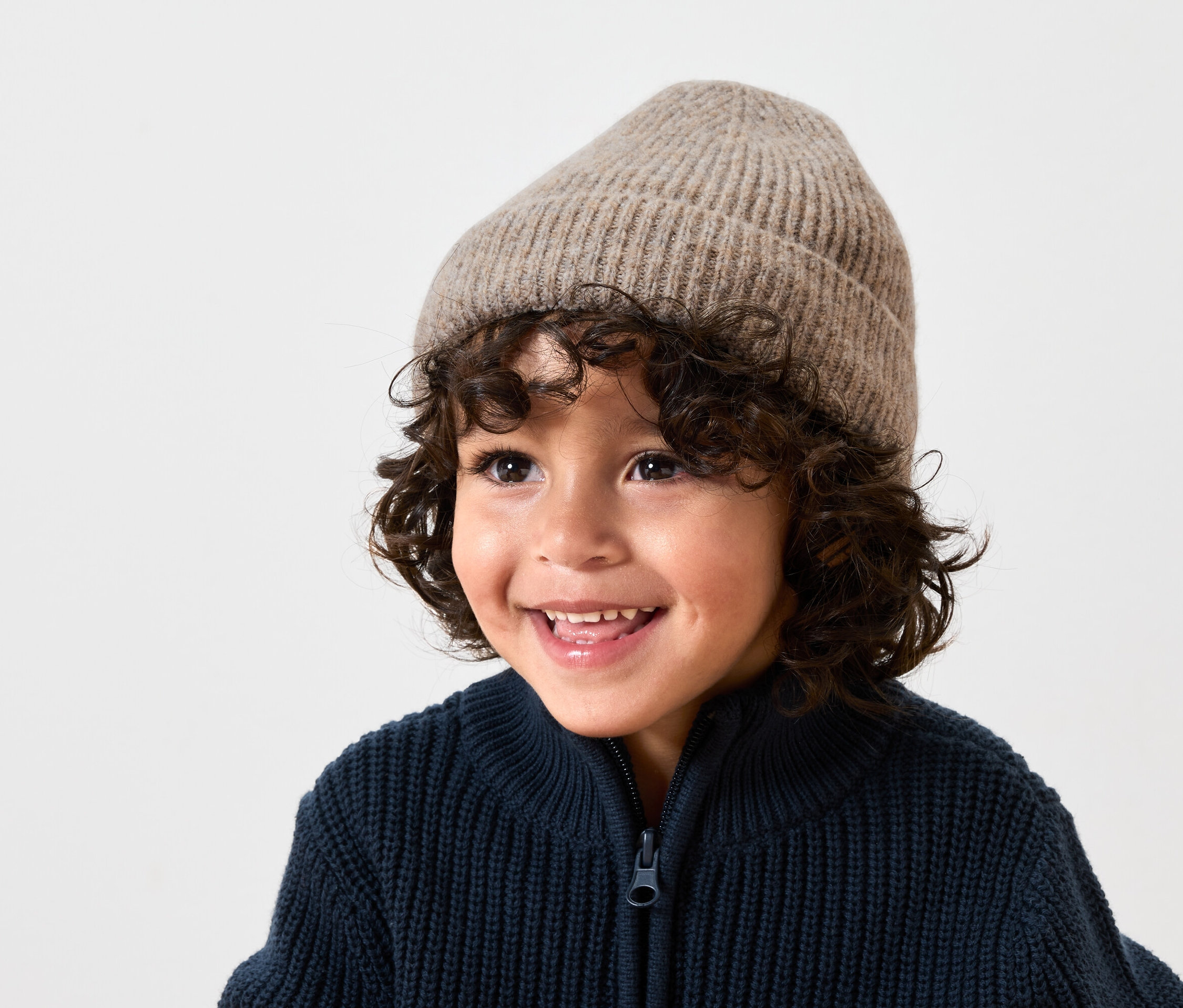 Uśmiechnięty chłopiec z brązowymi kręconymi włosami ma na sobie beżową wełnianą czapkę i granatowy sweter zapinany na zamek błyskawiczny.