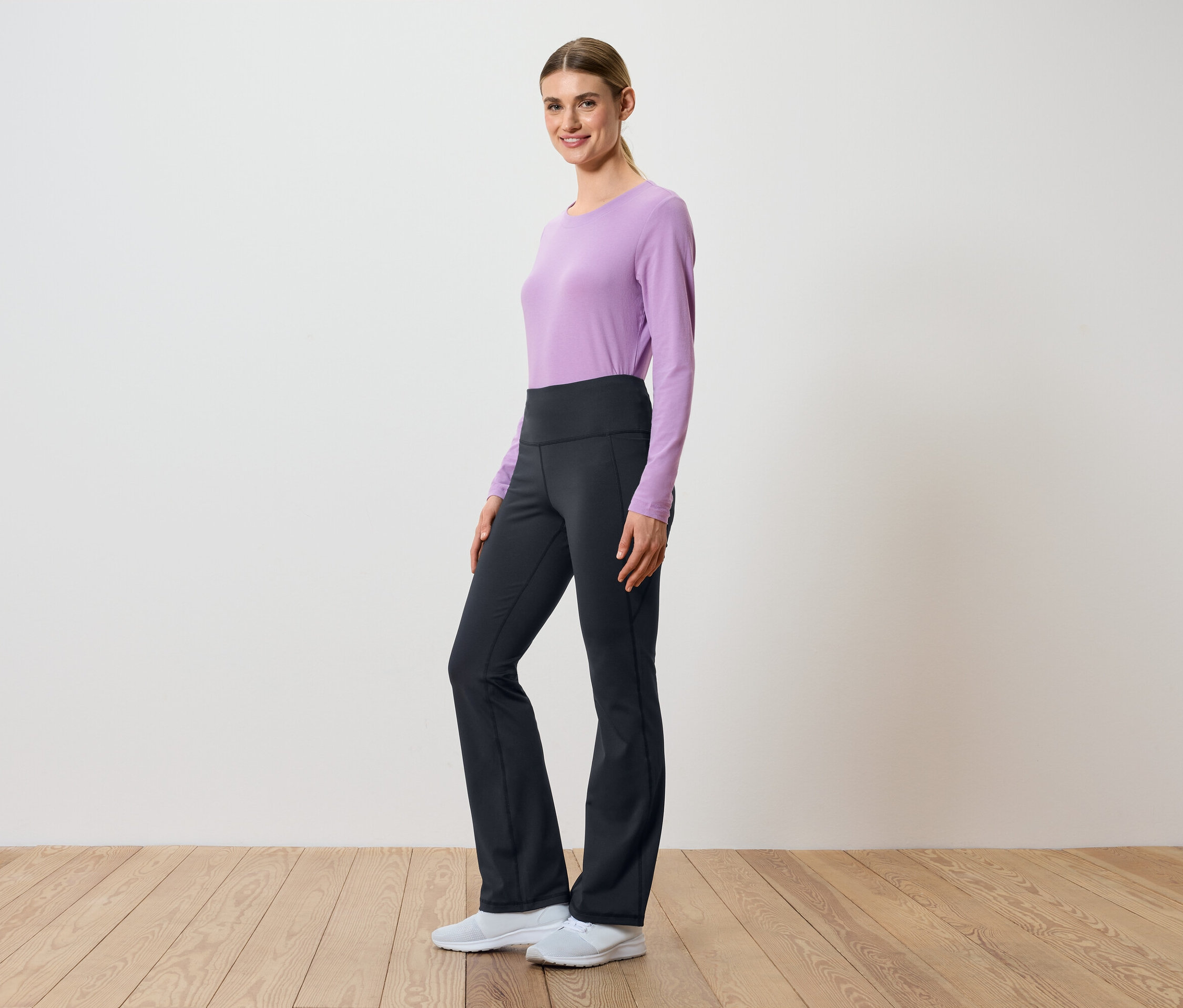 Kobieta pozuje bokiem w liliowej bluzce z długim rękawem i czarnych spodniach z wysokim stanem.