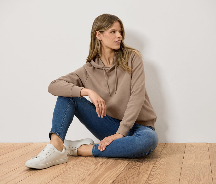 Kobieta siedzi na drewnianej podłodze, ubrana w brązową bluzę z kapturem, niebieskie dżinsy i białe tenisówki.