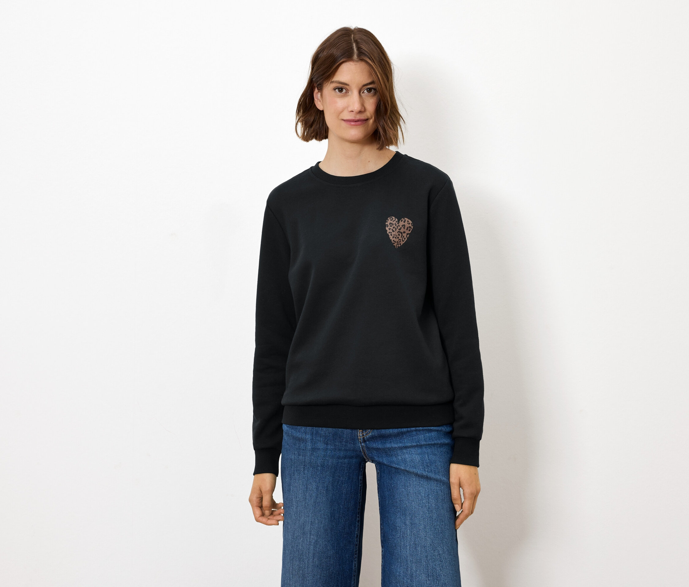 Kobieta stoi, ubrana w czarną bluzę z ozdobą w kształcie serca i dżinsy.