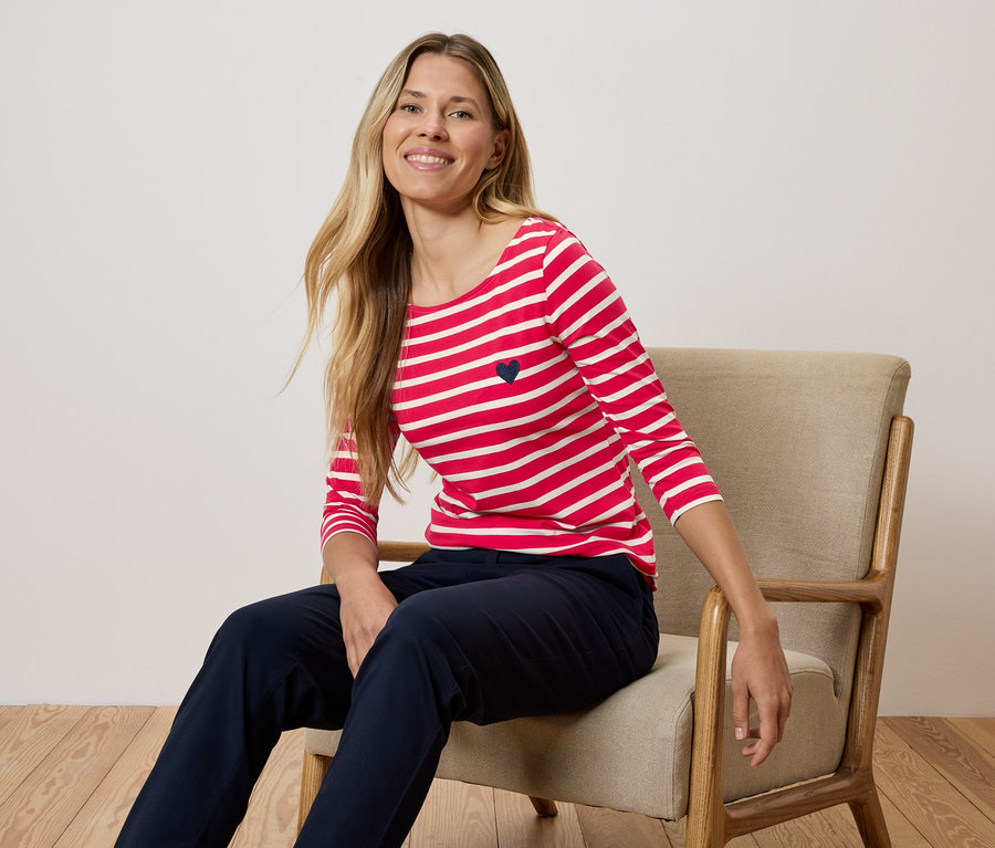 Kobieta siedzi na krześle, ubrana w czerwono-białą koszulkę w paski i granatowe spodnie.