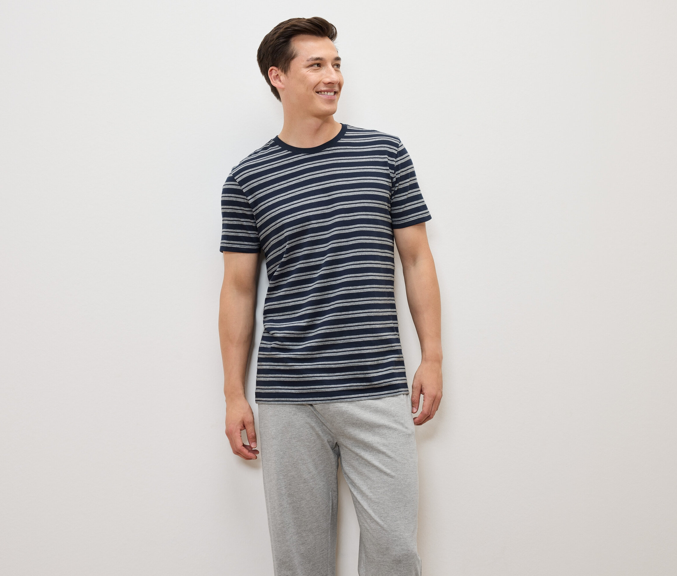Mężczyzna stoi, ubrany w niebiesko-szarą koszulkę w paski i szare spodnie.