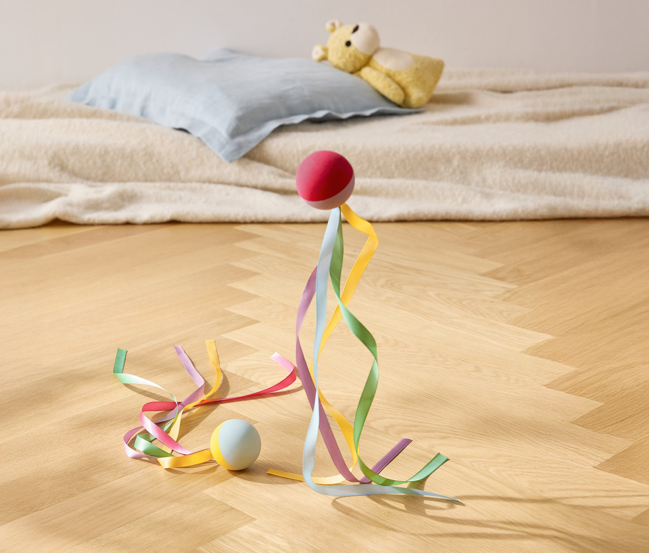 Czerwona piłka stoi z kolorowymi wstążkami na drewnianej podłodze, w tle poduszka i pluszowy miś.