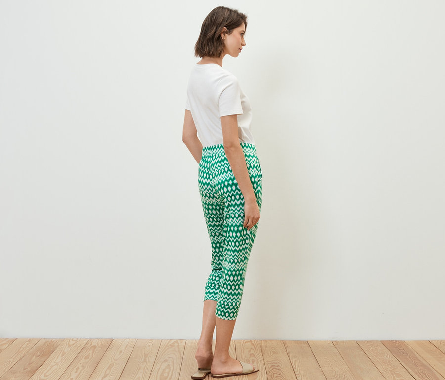 Kobieta stoi bokiem, ma na sobie białą koszulkę i wzorzyste spodnie dżersejowe o długości 3/4.