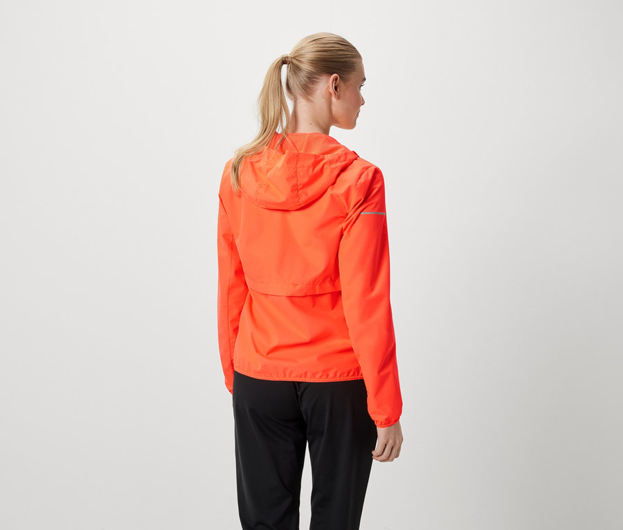 Kobieta z blond włosami związanymi w kucyk ma na sobie pomarańczową kurtkę i czarne spodnie.