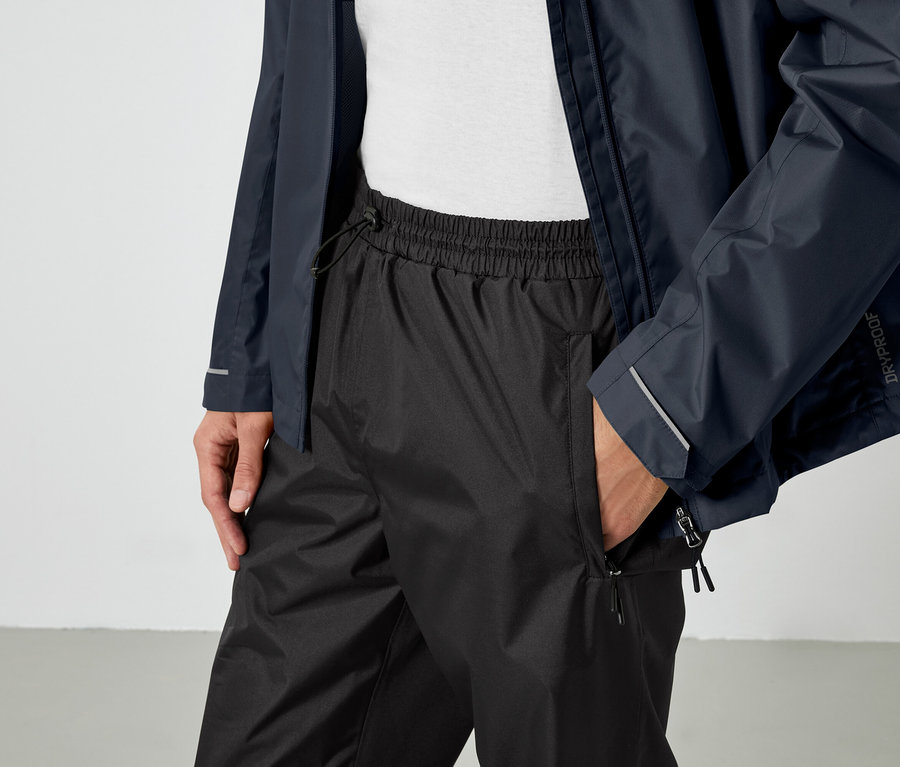 Mężczyzna trzyma rękę w kieszeni czarnych spodni i ma na sobie niebieską kurtkę.