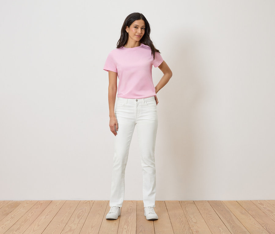 Kobieta stoi na tle białej ściany, ubrana w różowy T-shirt, białe dżinsy i białe tenisówki.