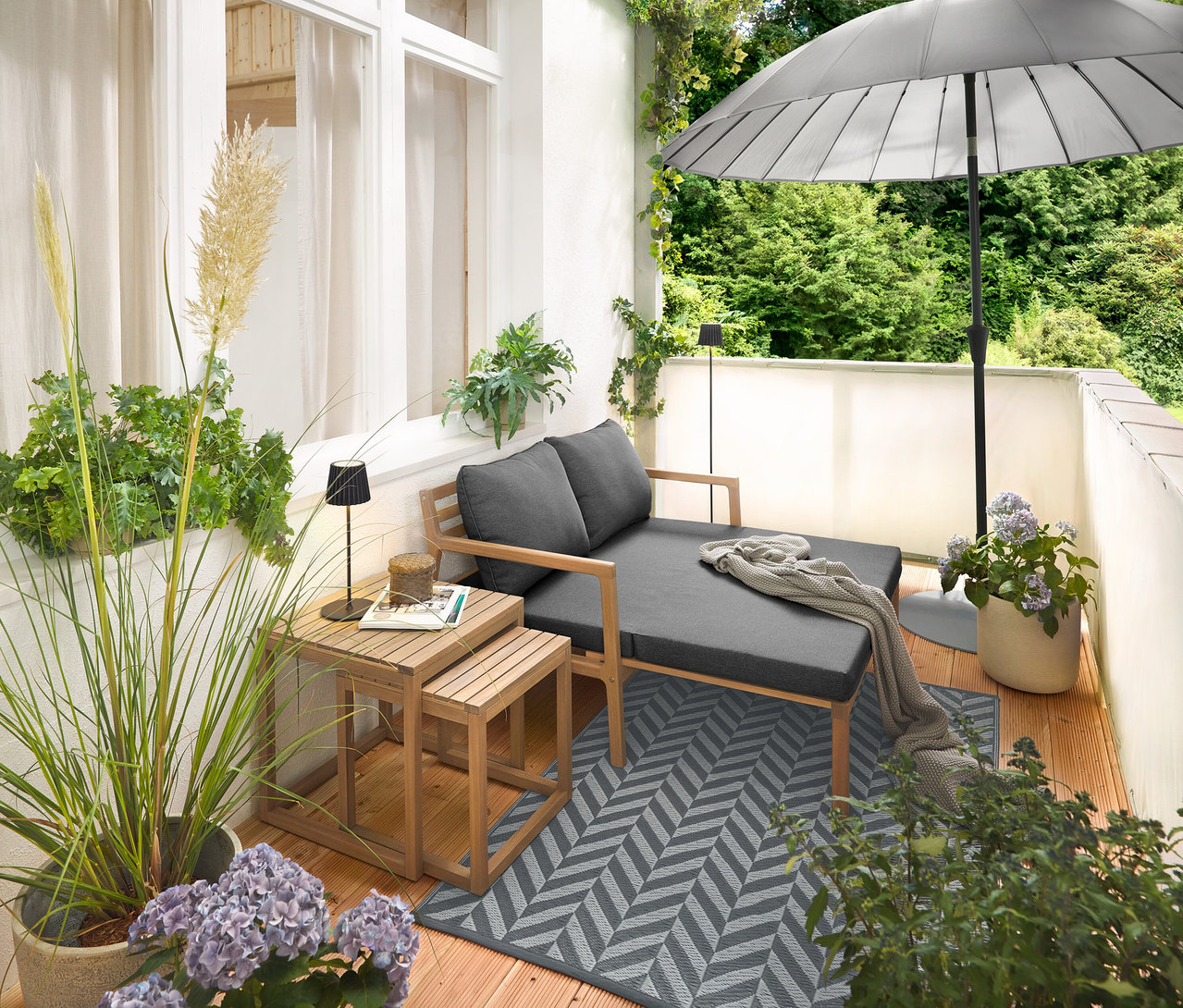 Aranżacja na balkonie z rozsuwaną sofą ogrodową, dywanem zewnętrznym (ok. 165 x 115 cm), parawanem balkonowym w kolorze kremowym, czarną akumulatorową lampą stołową LED do użytku na zewnątrz i czarną akumulatorową lampą stojącą LED do użytku na zewnątrz.