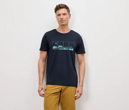 Mężczyzna stoi na tle ściany, ubrany w granatowy T-shirt z grafiką przedstawiającą cztery naturalne krajobrazy oraz khaki szorty.