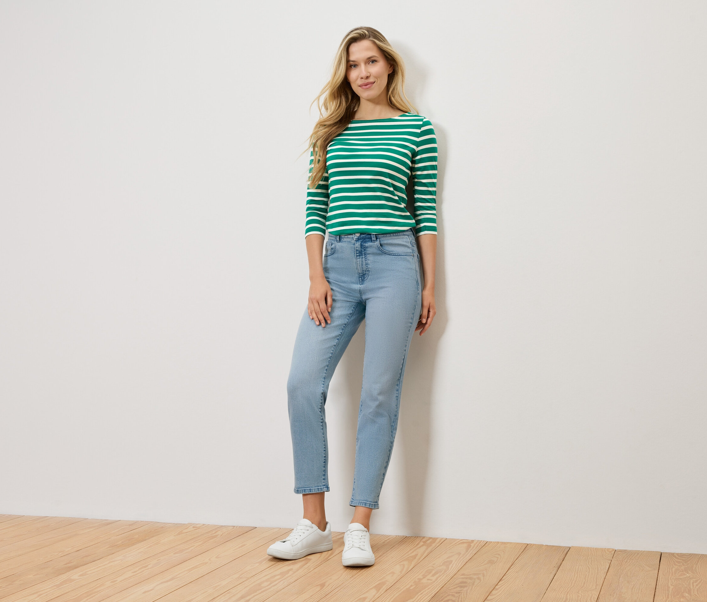 Blondynka stoi na tle ściany, ubrana w zielono-białą bluzkę z długim rękawem, jasnoniebieskie dżinsy i białe tenisówki.