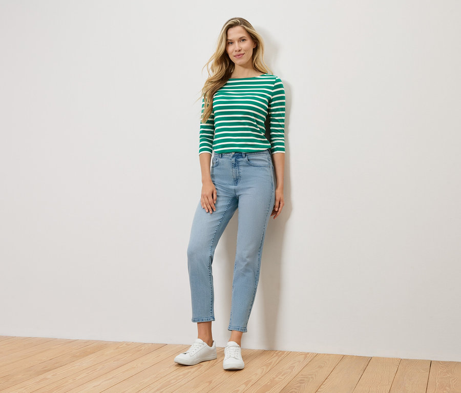 Blondynka stoi na tle ściany, ubrana w zielono-białą bluzkę z długim rękawem, jasnoniebieskie dżinsy i białe tenisówki.