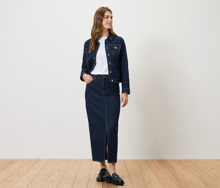 Kobieta stoi i ma na sobie dżinsową kurtkę i dżinsową spódnicę z rozcięciem.