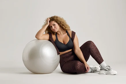 Kobieta o kręconych włosach siedzi obok srebrnej piłki gimnastycznej, ma na sobie czarny biustonosz sportowy, legginsy w zebrę i buty sportowe.
