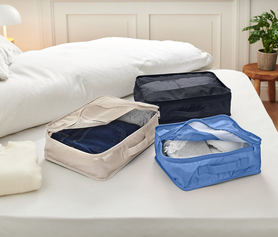 Na łóżku z białą pościelą leżą trzy otwarte kostki do pakowania. Kostki są beżowe, niebieskie i czarne i zawierają ubrania.