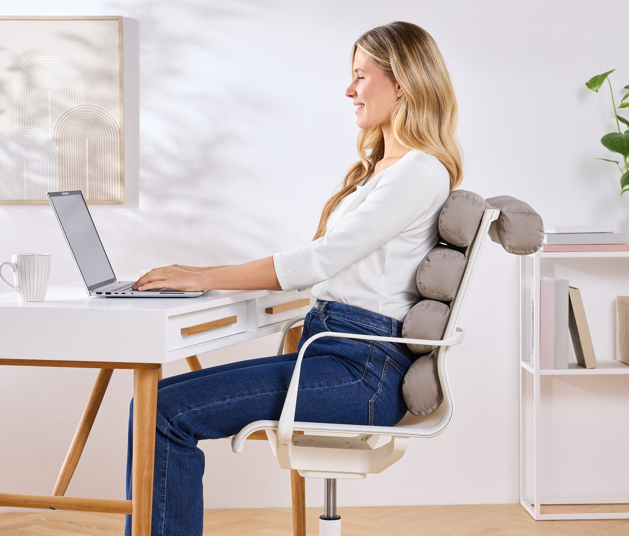 Kobieta siedzi przy biurku i korzysta z laptopa, z szarą poduszką pod plecy na krześle.