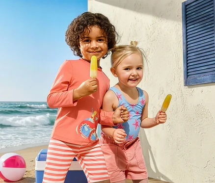 Dwoje dzieci z lodami na plaży. Dziewczynka ma na sobie strój kąpielowy Kinder-Badeanzug, Seesterne i dżinsowe szorty. Chłopiec nosi kombinezon Kinder-Kombi z ochroną UV i motywem ośmiornicy.