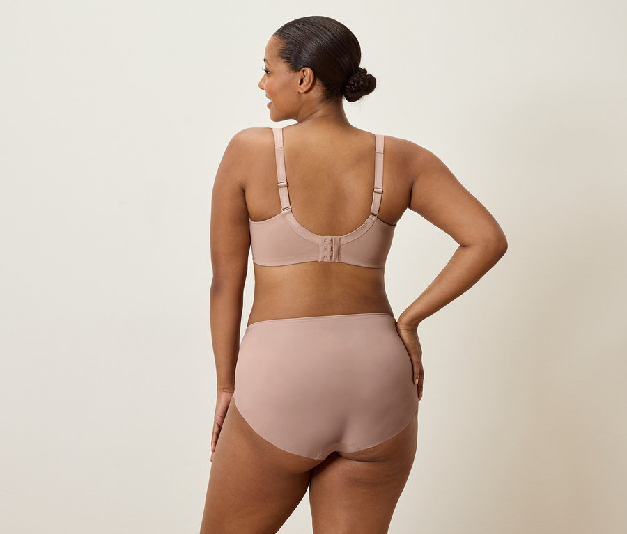Kobieta widziana od tyłu, ubrana w beżowy biustonosz i majtki, z jedną ręką na biodrze.