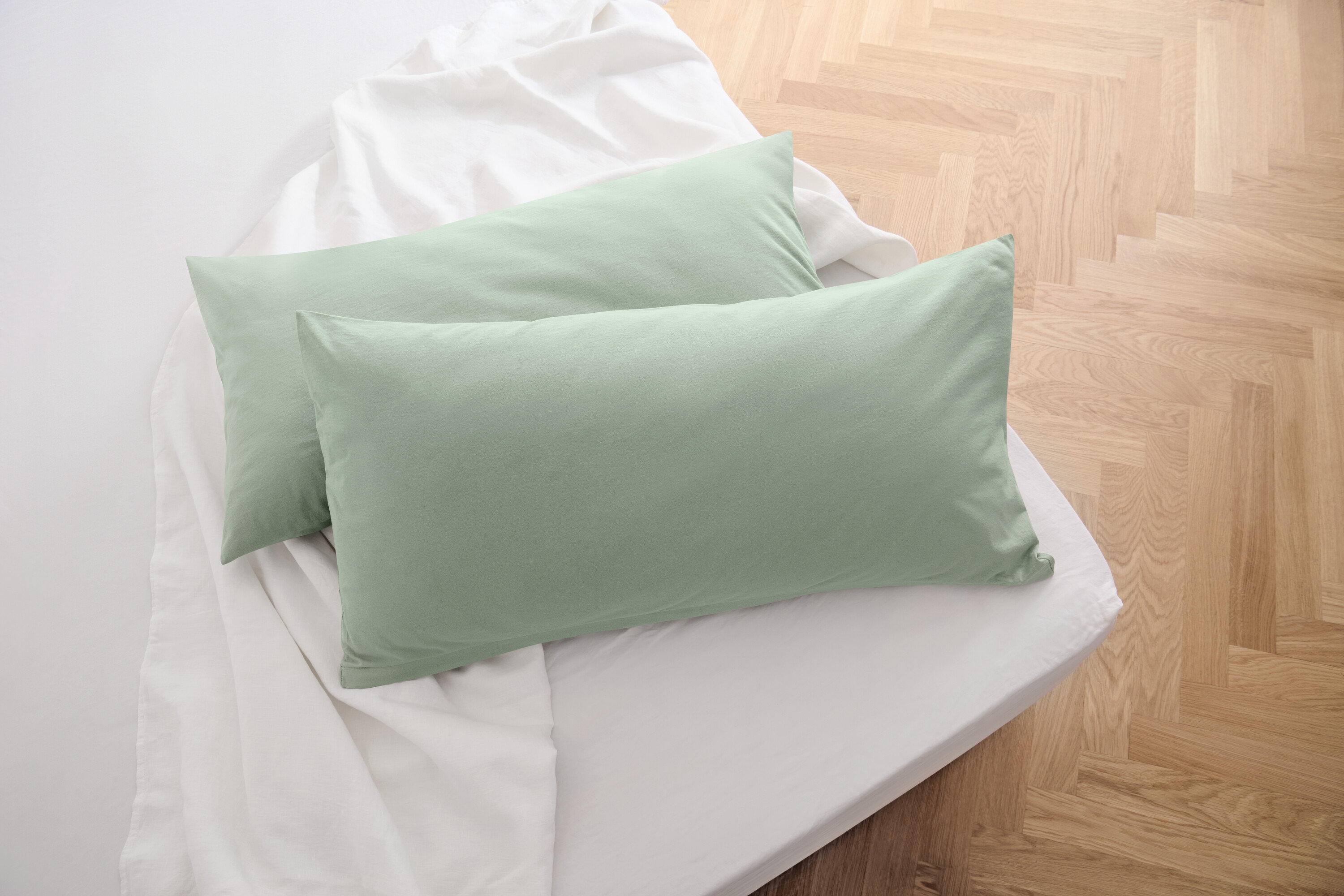 Dwie szałwiowe, dżersejowe poszewki na poduszki leżą na białym prześcieradle.