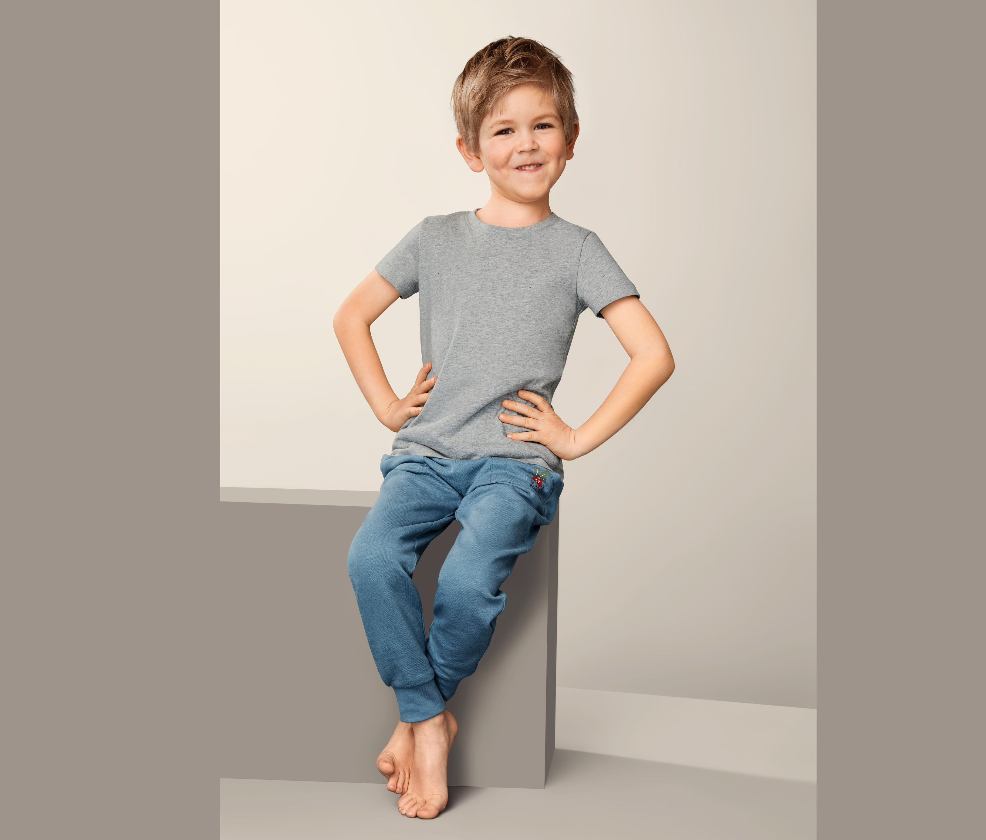 Chłopiec siedzi i ma na sobie szarą podkoszulkę dziecięcą (2 sztuki) i niebieskie spodnie dresowe.