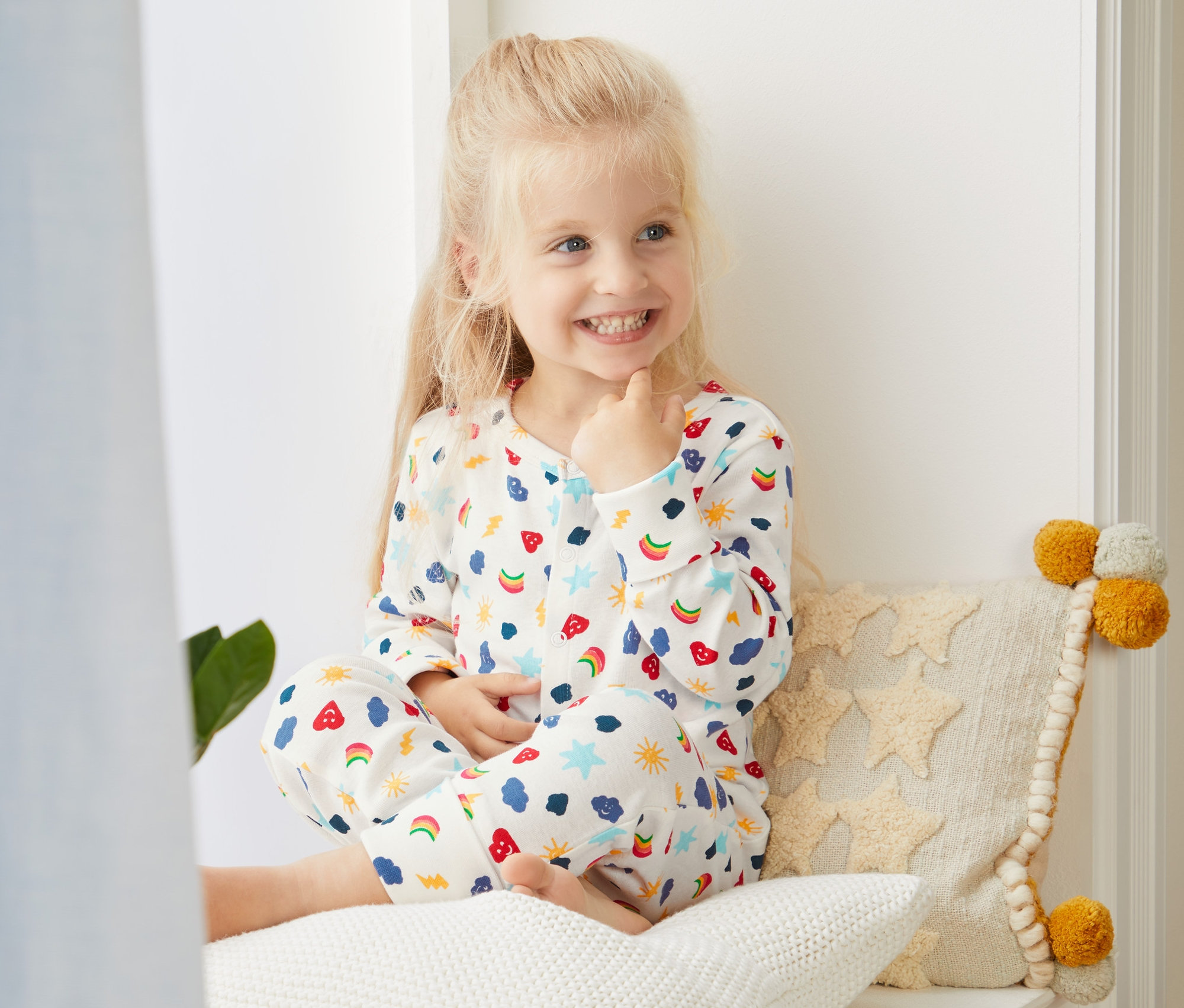 Uśmiechnięta blondwłosa dziewczynka siedzi po turecku na parapecie w piżamie z bawełny ekologicznej w białe wzory.