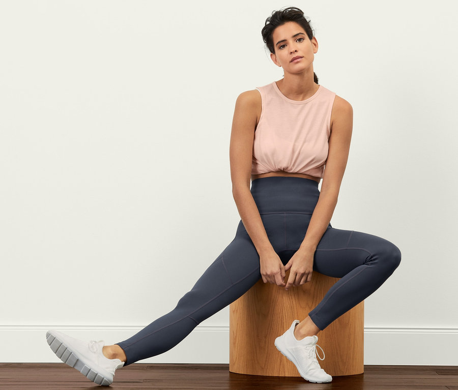 Kobieta siedzi na drewnianym stołku, ma na sobie legginsy sportowe »Sculpture«, top i tenisówki.