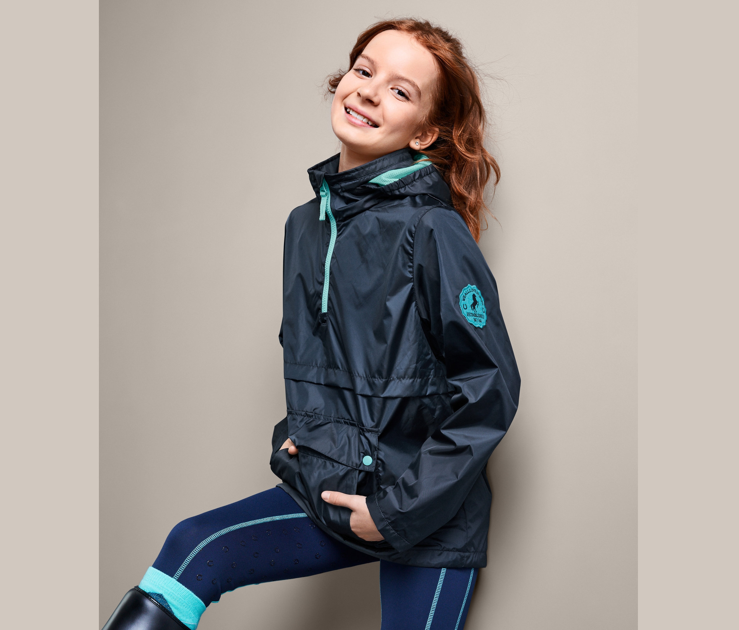 Uśmiechnięta nastolatka pozuje w kurtce przeciwdeszczowej zakładanej przez głowę, granatowych legginsach i czarnych butach do jazdy konnej.