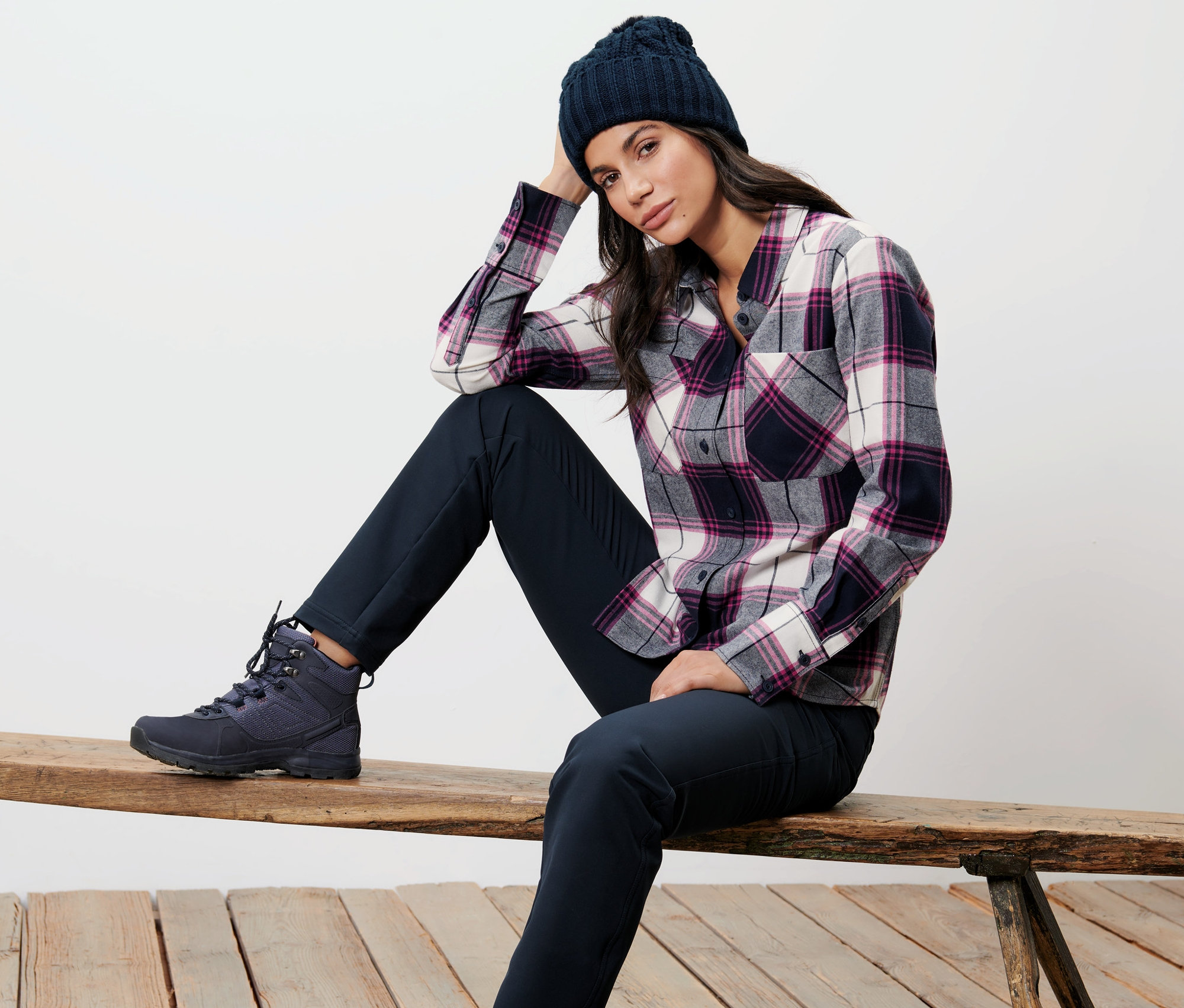 Kobieta siedzi na ławce w koszuli w kratę i czapce.