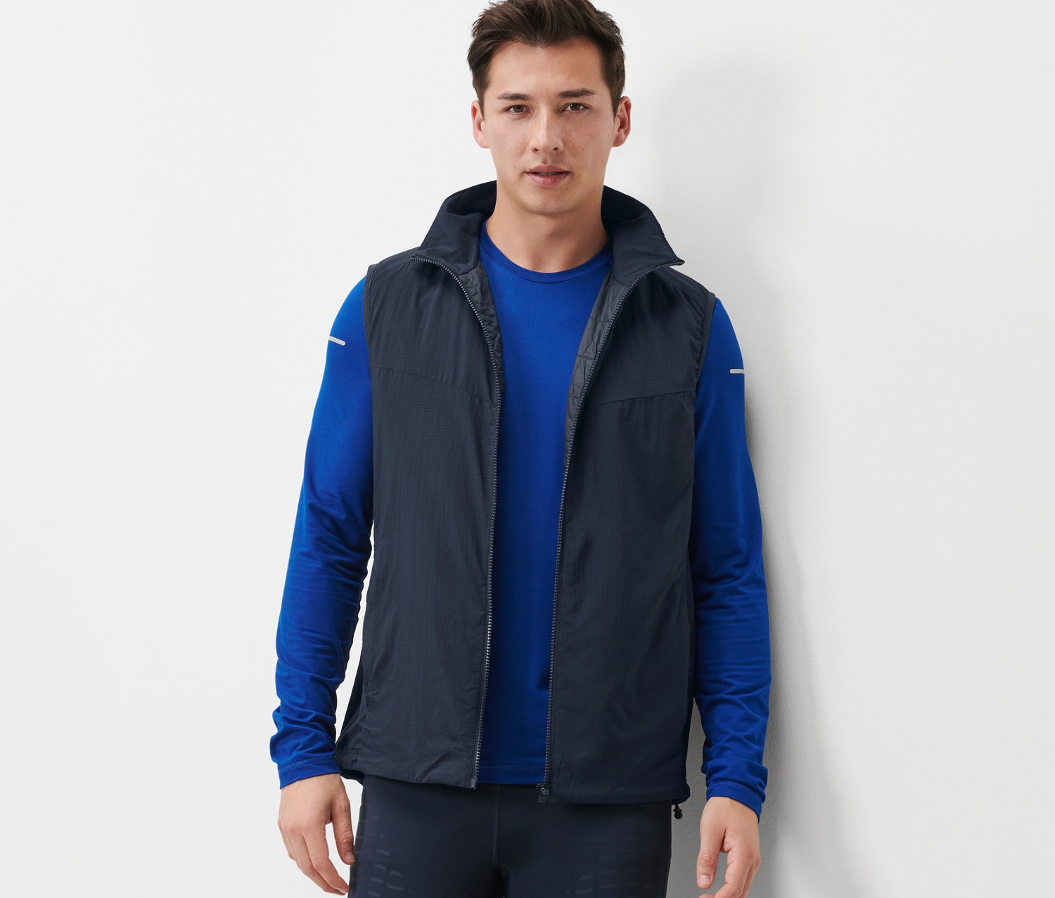 Mężczyzna ma na sobie kamizelkę funkcyjną, niebieską koszulkę z długim rękawem i ciemne legginsy.