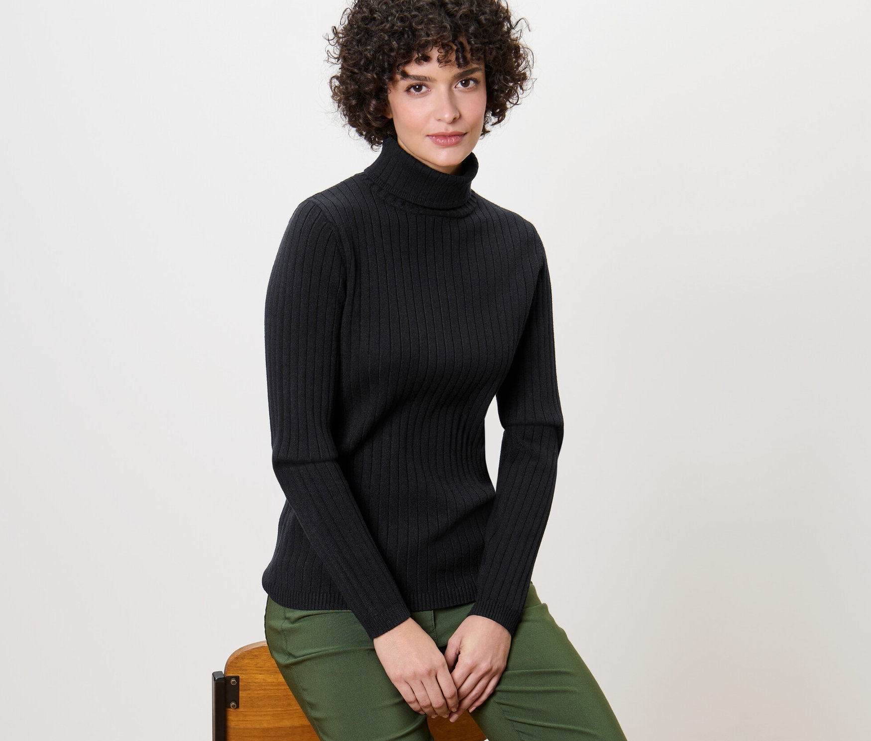 Kobieta siedzi na krześle, ubrana w czarny golf z dzianiny o splocie ściągaczowym.