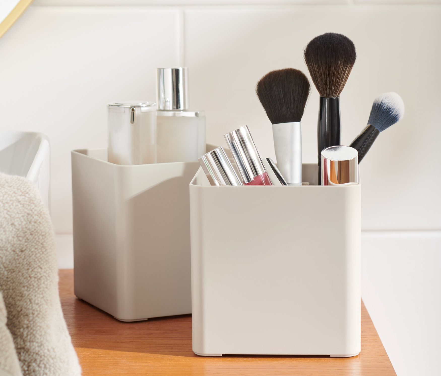 Dwa beżowe Organizery, 2 sztuki na drewnianej szafce. W jednym z nich znajdują się pędzle do makijażu i szminki.