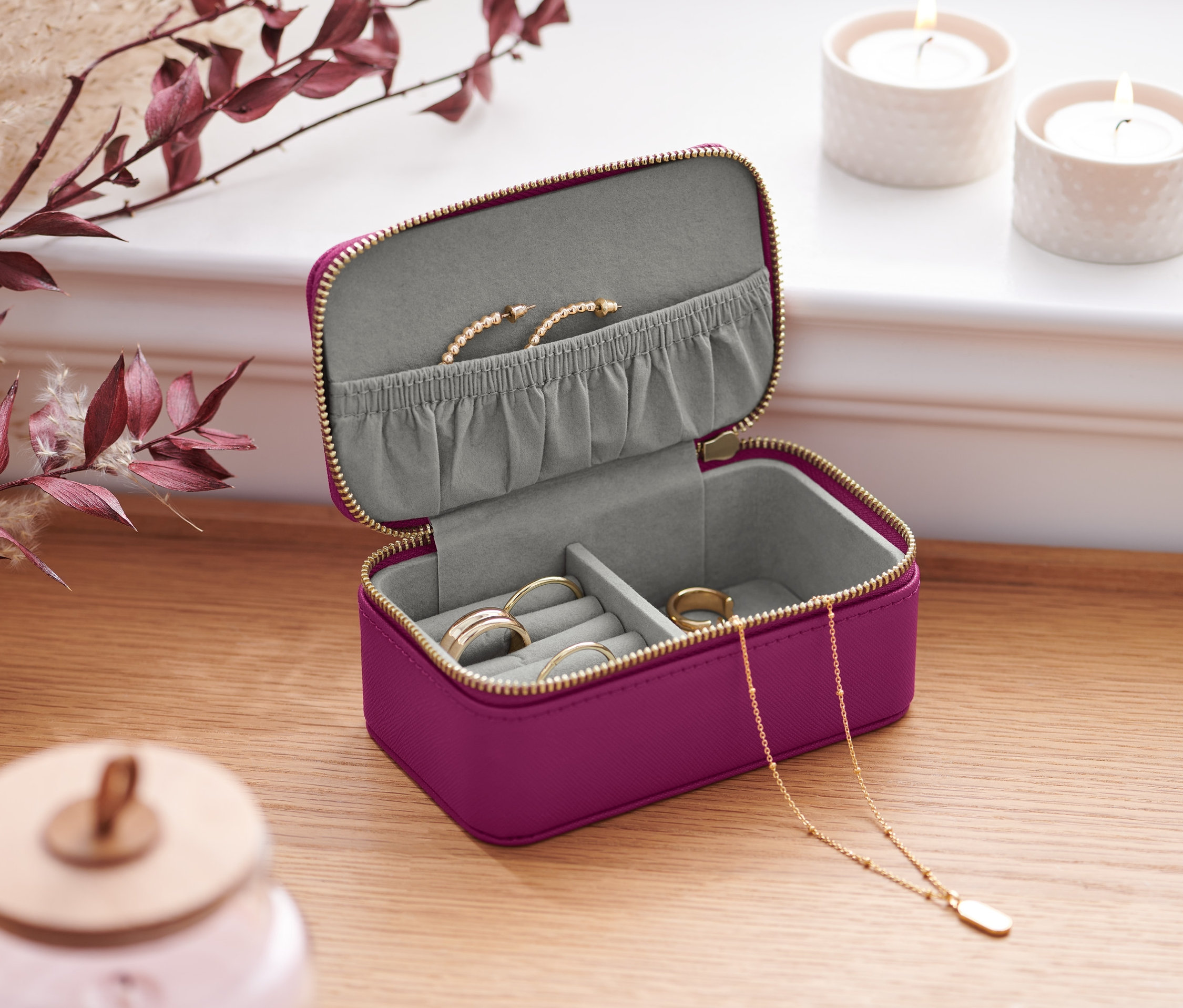 Otwarta mała kasetka na biżuterię w kolorze fioletowym ze złotym zamkiem błyskawicznym na drewnianym stole. W środku znajdują się pierścionki, naszyjnik i bransoletka.