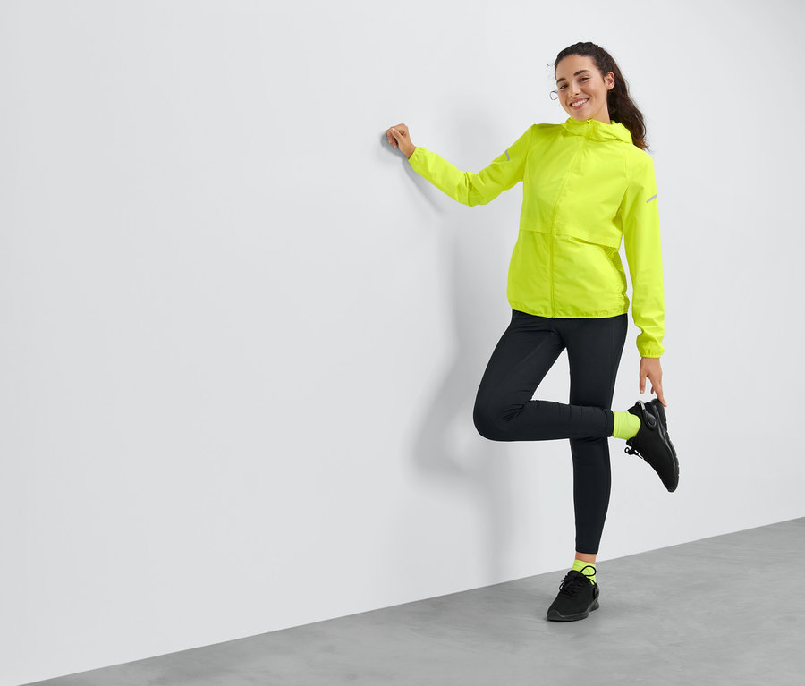 Kobieta opiera się o ścianę, pozując w żółtej wiatroszczelnej kurtce do biegania.