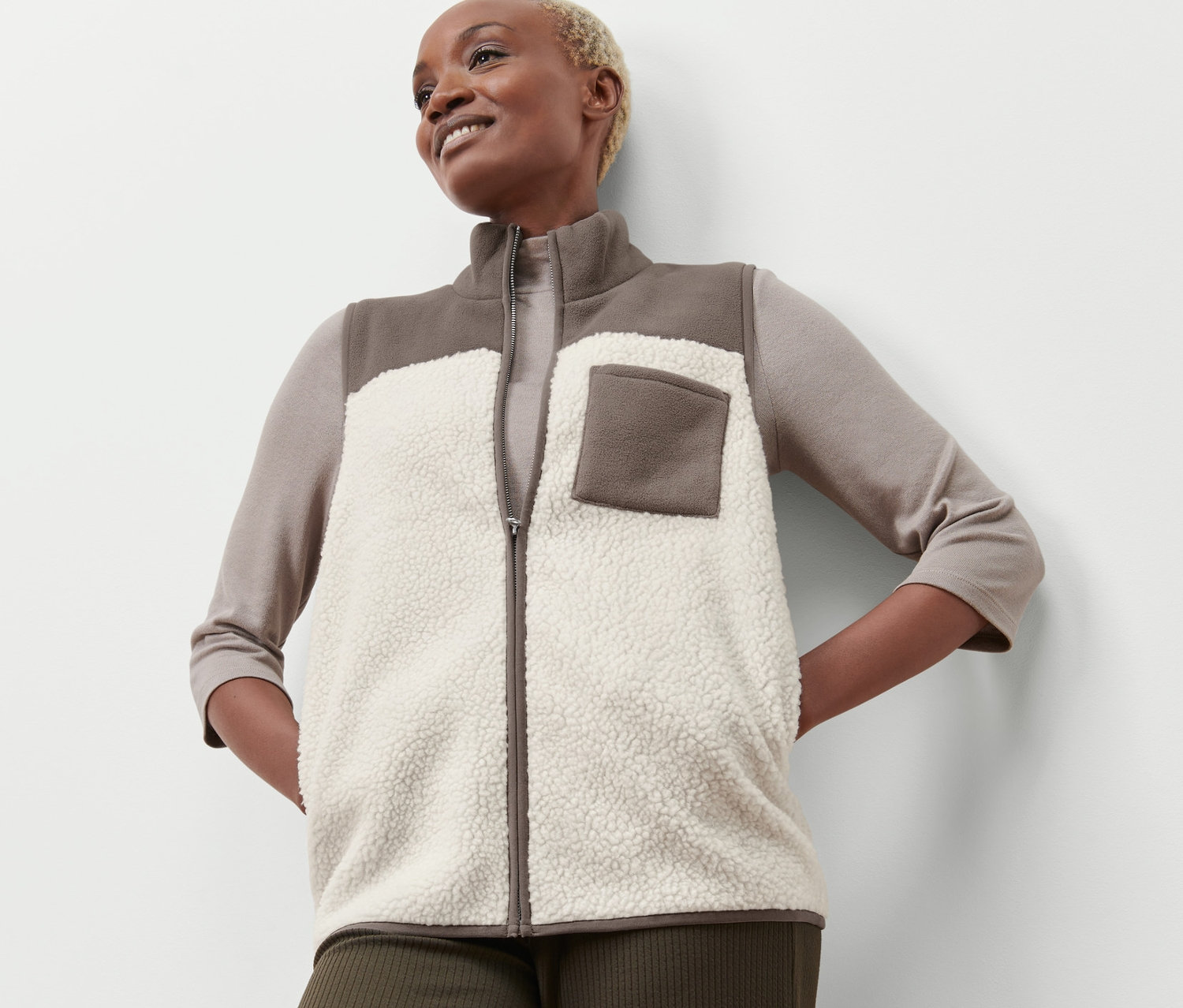 Kobieta z krótkimi blond włosami pozuje w beżowo-brązowej pluszowej kamizelce.