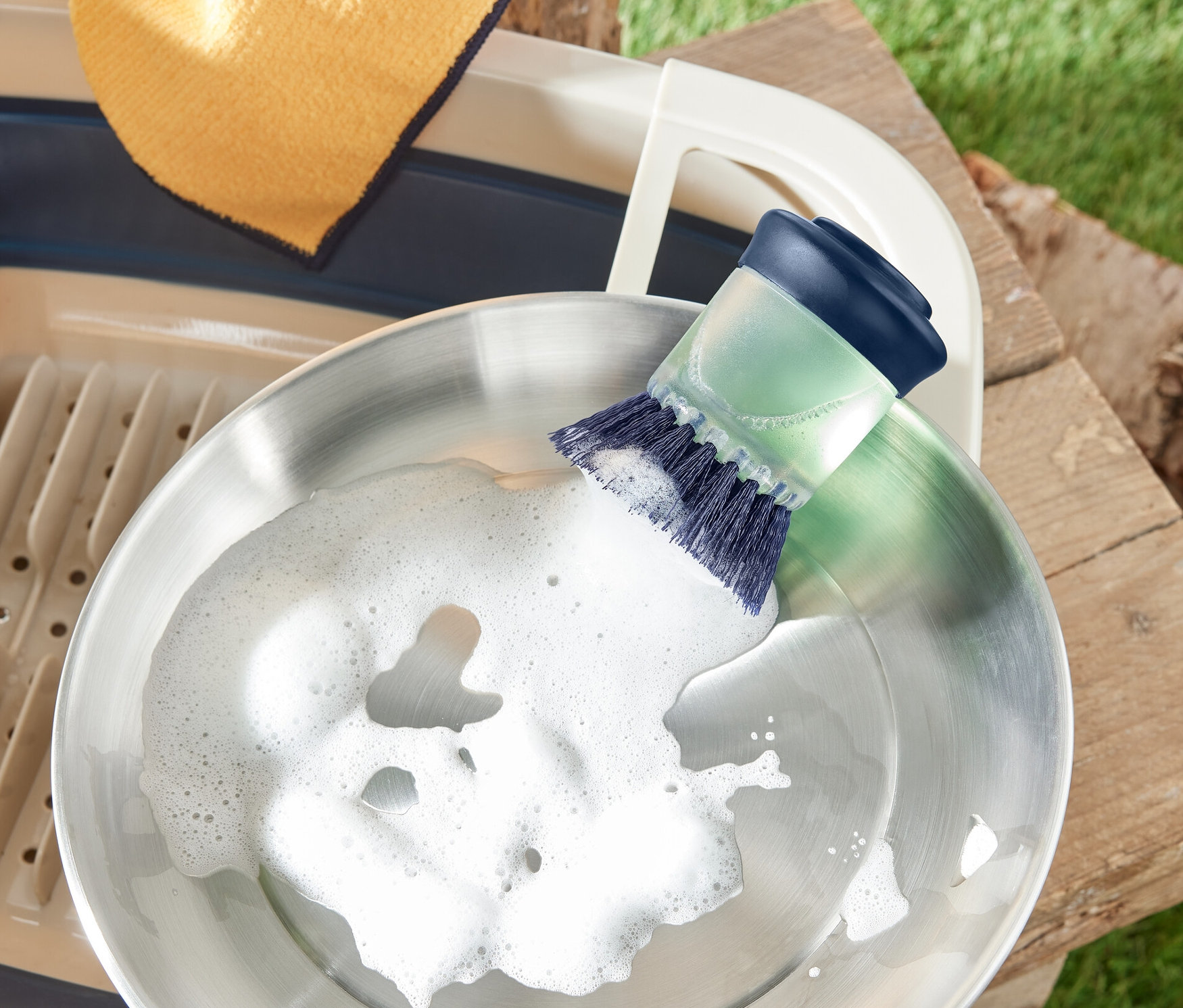 Szczotka do mycia naczyń z dozownikiem myje stalową patelnię. Obok ociekacz i ścierka.